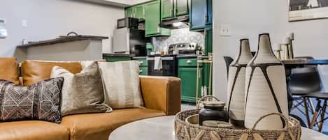 A living room with a brown leather sofa, a round table, and a kitchen in the background.