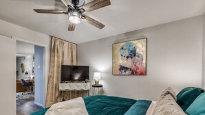 Un dormitorio con cama, ventilador de techo, televisor, escritorio y un cuadro.