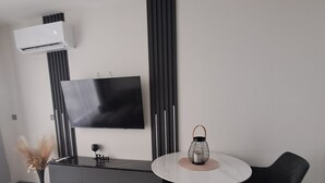 Una sala moderna con una mesa de comedor redonda, dos sillas, un televisor de pantalla plana en la pared, una consola negra y un florero con flores secas.