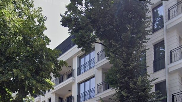 Un edificio residencial de varios pisos con balcones, ventanas grandes y un árbol en la fachada.