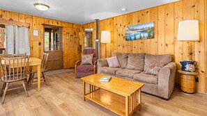 A wooden-paneled room with a sofa, a coffee table, a dining table with chairs, and a painting on the wall. - Charming 2-bedroom, 2-bathroom Machin Cottage #1 (Estes Park)