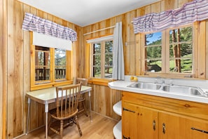 A wooden cabin interior with a dining table, chairs, a kitchen with a sink, and large windows.