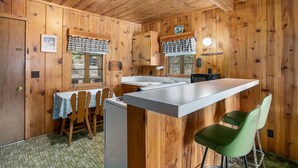 Interior de una cabaña de madera con una zona de cocina que incluye una barra, armarios de madera y una mesa de comedor con sillas.