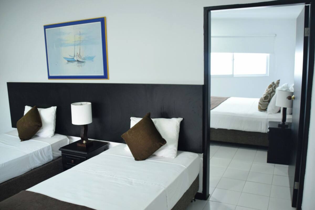 Habitación de hotel con dos camas, una mesita de noche con lámpara, un cuadro enmarcado en la pared y un espejo que refleja la habitación.