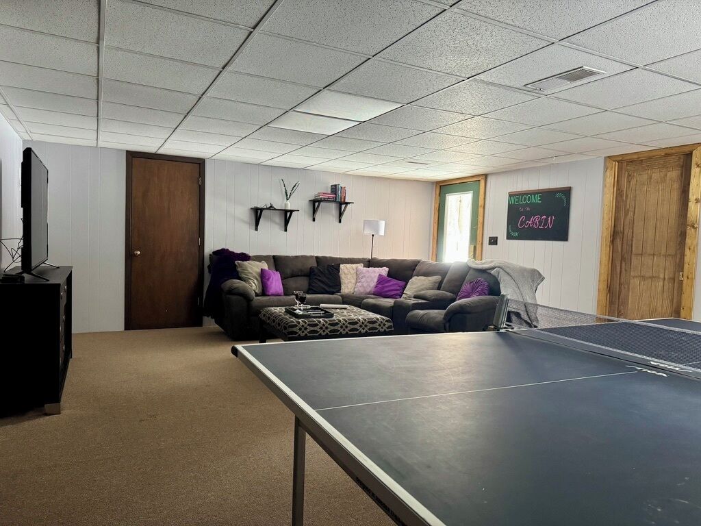 Una habitación con mesa de ping pong, un sofá con cojines, una televisión y un cartel de bienvenida en la pared.