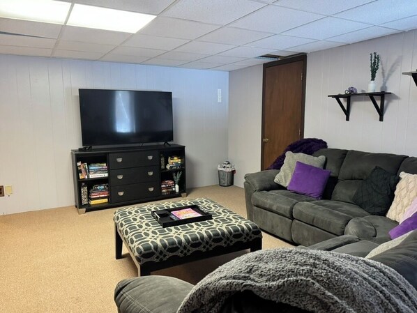 A living room with a flat-screen TV, a bookshelf, a grey sofa with cushions, and a patterned ottoman.