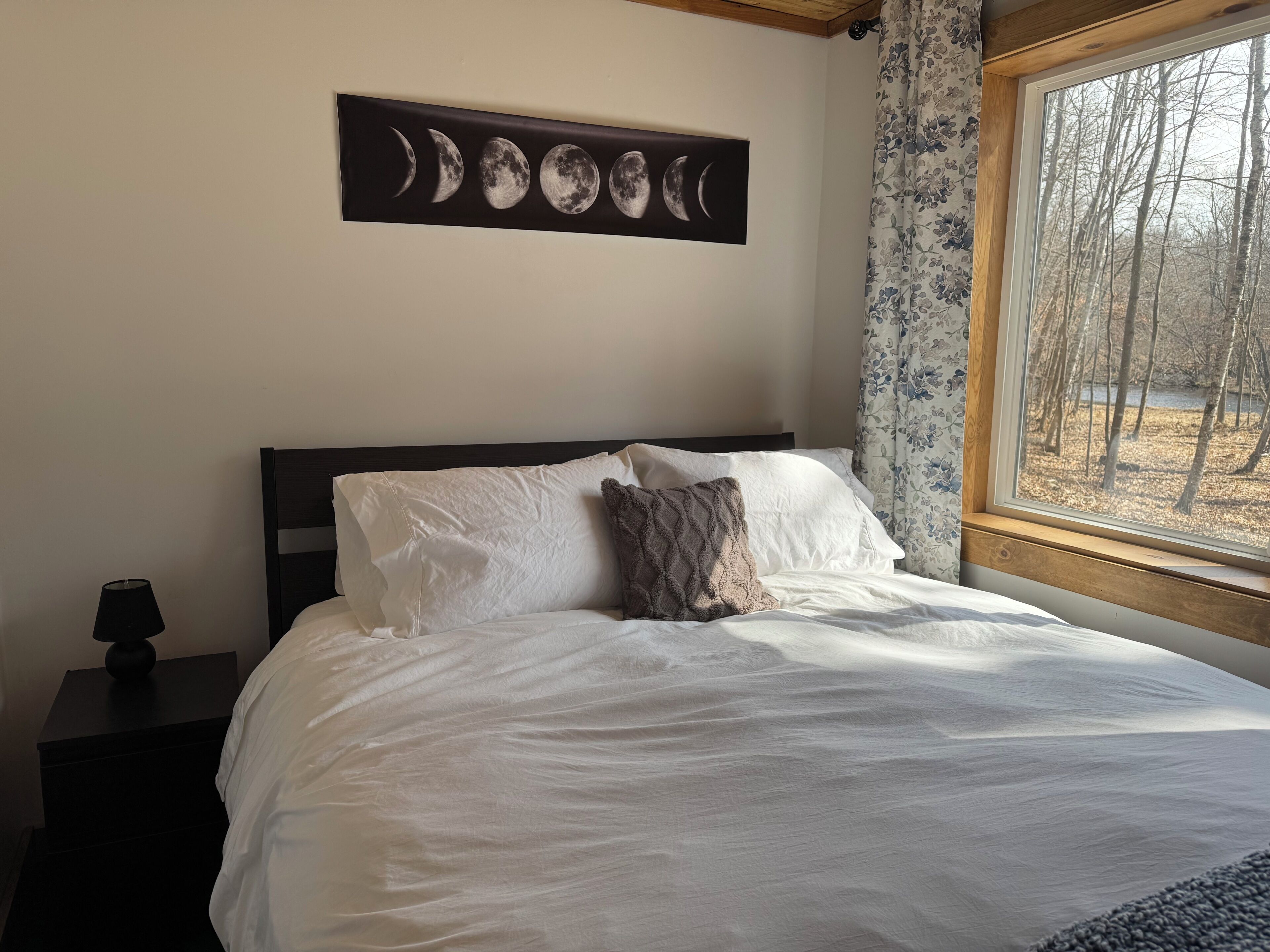 A bedroom with a bed, pillows, a nightstand, a lamp, a wall-mounted moon phase art piece, and a window with a view of trees.