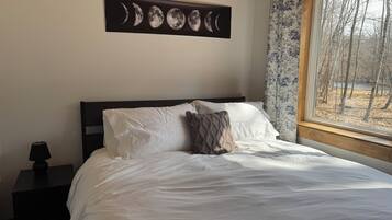 A bedroom with a bed, pillows, a nightstand, a lamp, a wall-mounted moon phase art piece, and a window with a view of trees.