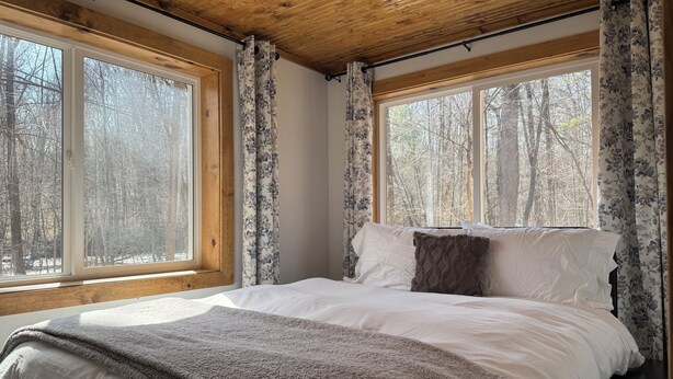 Une chambre avec un plafond en bois, de grandes fenêtres avec des rideaux, un lit avec une literie blanche et une couverture.