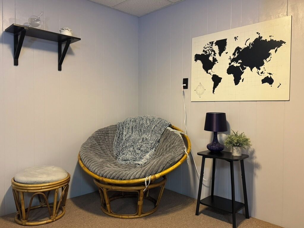 Habitación de esquina con una silla de ratán, un taburete pequeño, un mapa del mundo en la pared, una lámpara y una estantería con adornos.