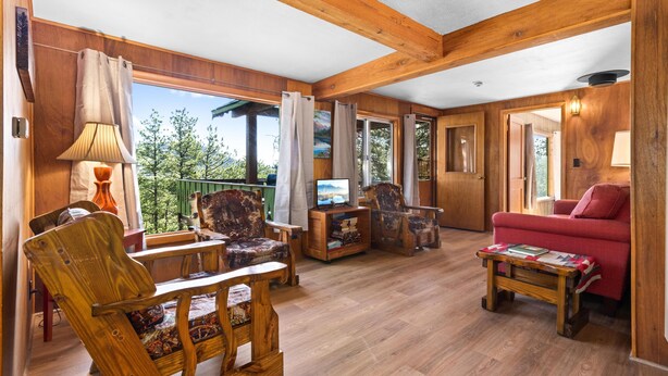 Ein Wohnbereich mit Holzboden, einem roten Sofa, Holzstühlen, einem Fernseher und Blick ins Freie.