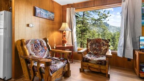 Ein Holzinnenraum mit zwei gemusterten Stühlen, einem Tisch, einer Lampe und Blick auf einen Wald durch ein Fenster.
