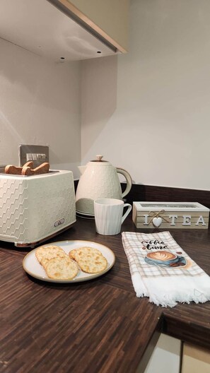 Uma bancada de cozinha com uma torradeira, uma cafeteira, uma chávena, um prato de bolachas e uma caixa com tema de chá.