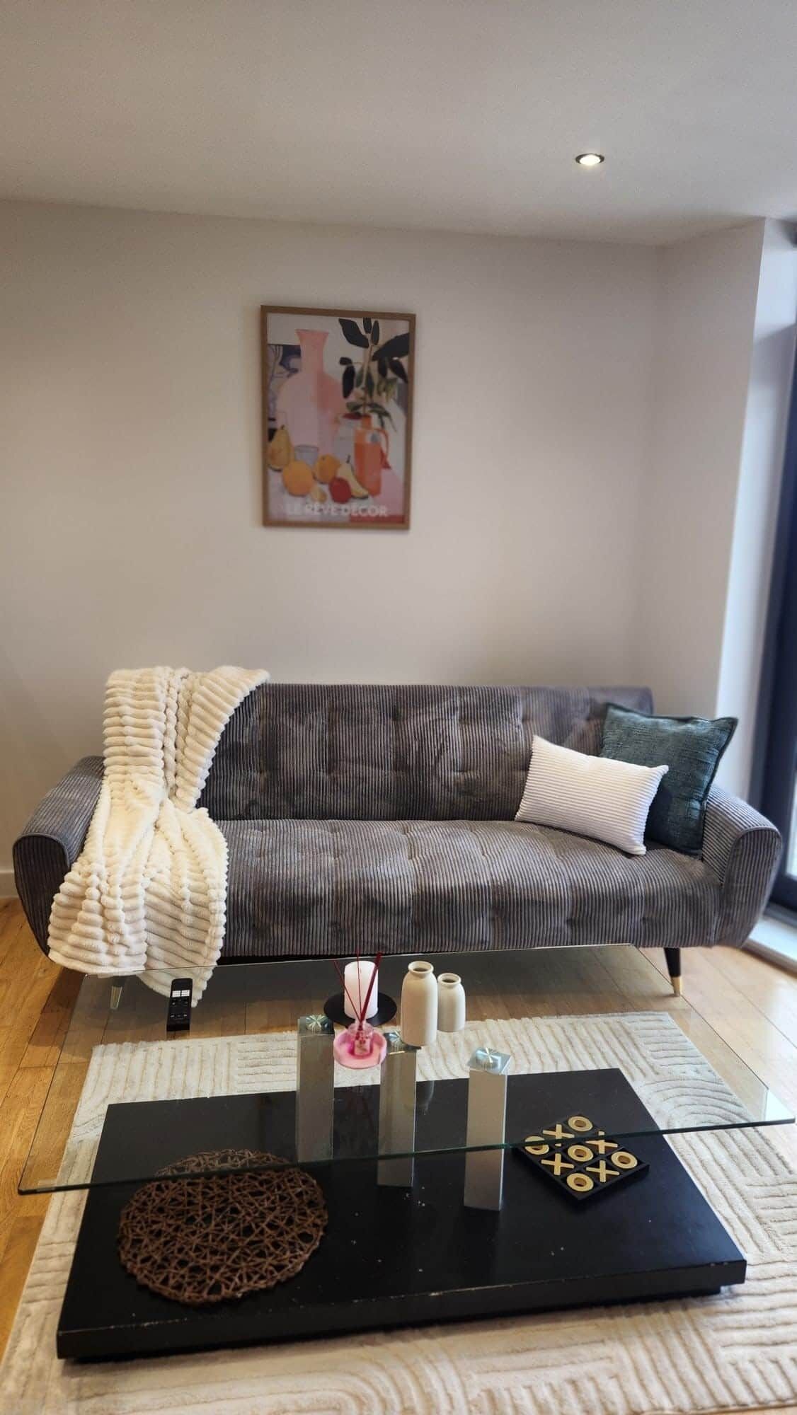 Un soggiorno moderno con un divano grigio, una coperta bianca, un tavolino da caffè in vetro e oggetti decorativi, e un quadro appeso al muro.