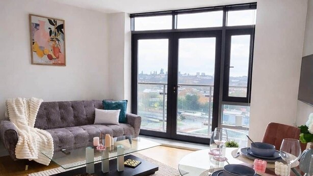 Una sala de estar moderna con un sofá, una mesa de centro de vidrio, una mesa de comedor para dos personas y un amplio ventanal con vista a la ciudad.