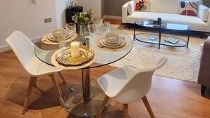 A dining area with a glass table, white chairs, and a white sofa in the background. - Urban Retreat | Leeds City Centre | Sleeps 4 (Leeds)