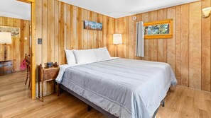 A wooden-paneled room with a bed, a nightstand, a lamp, and a painting on the wall.