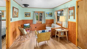 A cozy room with wooden walls and floors, a rocking chair, a bed, a desk with a lamp, and framed pictures on the walls. - Machin's Cottage #7 is calling! Pet Friendly (Estes Park)