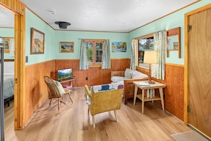 Una habitación acogedora con paredes y suelos de madera, un sillón mecedor, una cama, un escritorio con lámpara y cuadros en las paredes.