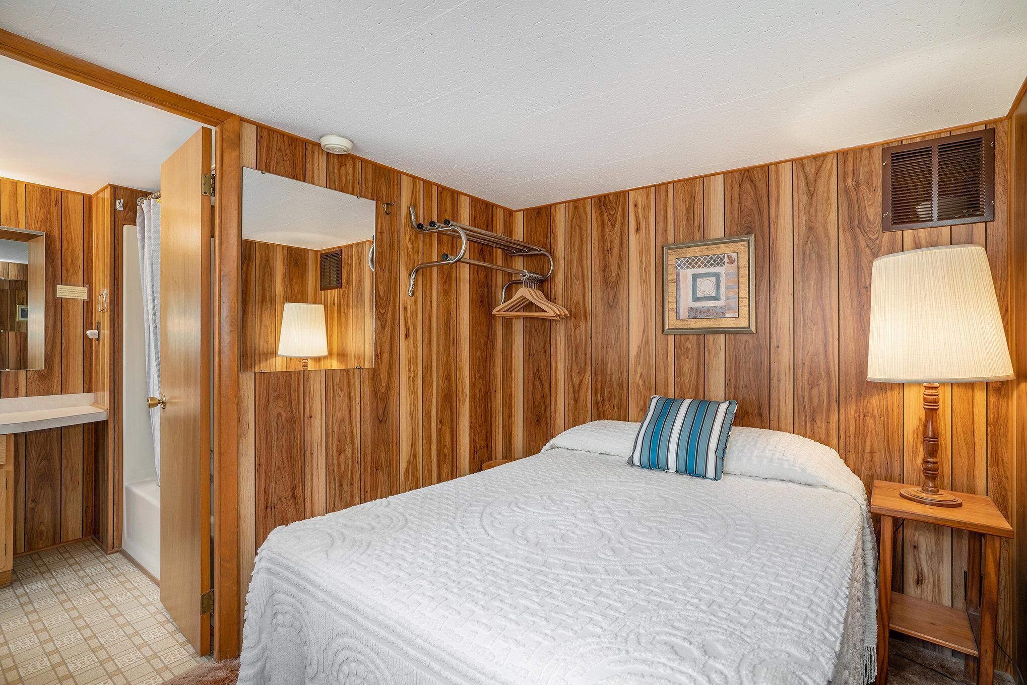 Slaapkamer met een wand van houten panelen, een bed met een witte quilt, een nachtkastje met een lamp, een wandrek voor handdoeken en een lijst met een afbeelding.