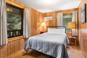 A wooden-paneled room with a bed, two bedside lamps, a window with curtains, and a wall-mounted picture.