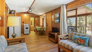 A cozy cabin interior with wooden walls, a dining area, a kitchen with a stove, and a sofa with a camping-themed pillow.