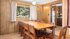 A wooden-paneled room with a large dining table, chairs, a window with curtains, and a kitchen area visible through an open door.
