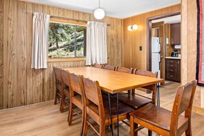 A wooden-paneled room with a large dining table, chairs, a window with curtains, and a kitchen area visible through an open door. - Escape to the embrace of nature at Machin's Cottage #15 Pet Friendly (Estes Park)