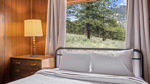 A bedroom with a wooden headboard, a bedside table with a lamp, a window with curtains, and a view of a forested hillside.