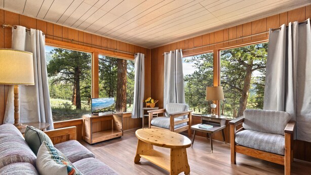 A cozy room with wooden walls and floors, a sofa, two chairs, a small wooden table, and large windows with curtains.
