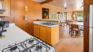 A kitchen with wooden cabinets, a central island with a sink, a dining area with wooden tables and chairs, and a view of greenery outside.