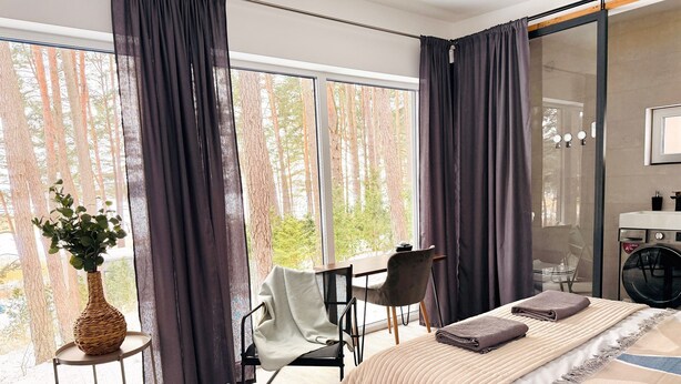 A modern bedroom with a large window, a bed with a beige blanket, a chair, a small table with a plant, and a washing machine.