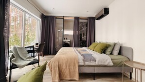 A modern bedroom with a bed, a sofa, a chair, and a small table. There is a window with curtains, a ceiling fan, and a wall-mounted air conditioning unit.