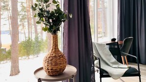 A room with a large window, a wicker vase with green foliage, a chair with a green cushion, and a table with a bowl.