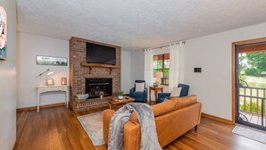 A living room with a brick fireplace, a sofa, and a television mounted above it. - Smoky Mountain Hideaway | Spacious Yard & Firepit (Maryville)