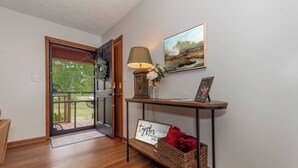 A wooden-floored hallway with a door leading to a balcony, a table with a lamp, a framed picture, and a sign on the wall. - Smoky Mountain Hideaway | Spacious Yard & Firepit (Maryville)