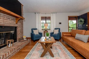 Un salon avec une cheminée en briques, deux fauteuils bleus, une table basse en bois et un canapé marron.