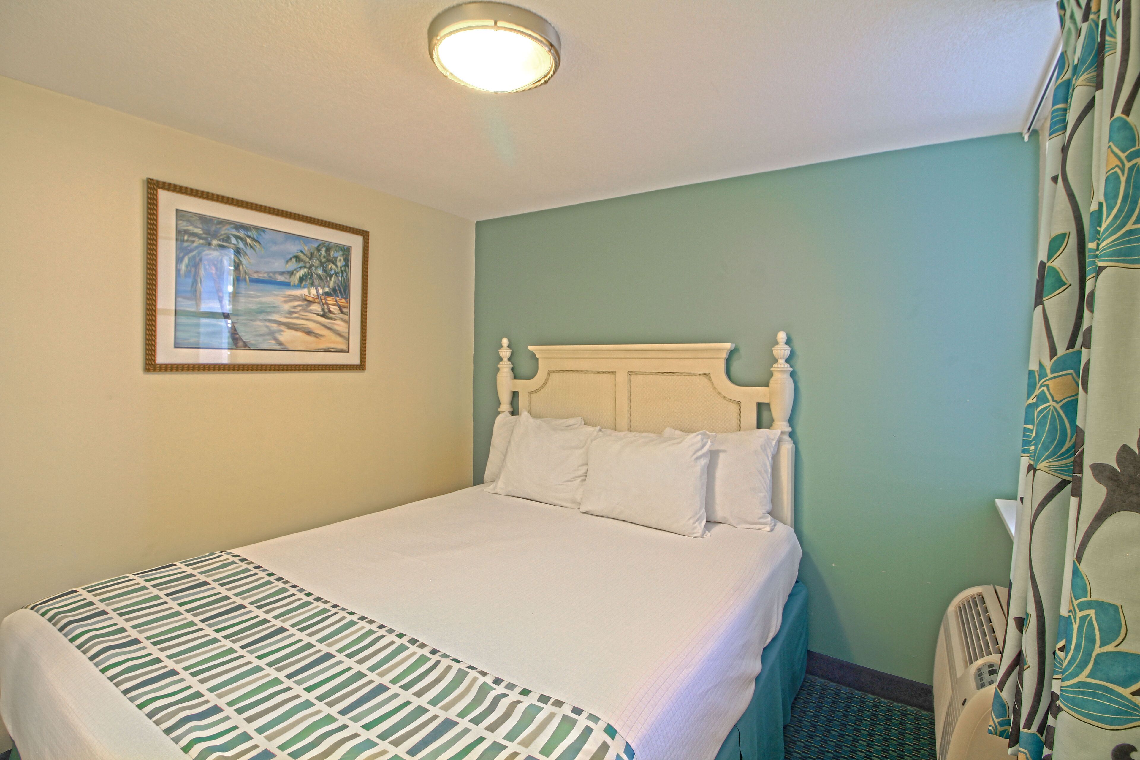A neatly made bed with a patterned bedspread, a white headboard, and a framed picture of a beach scene on the wall.
