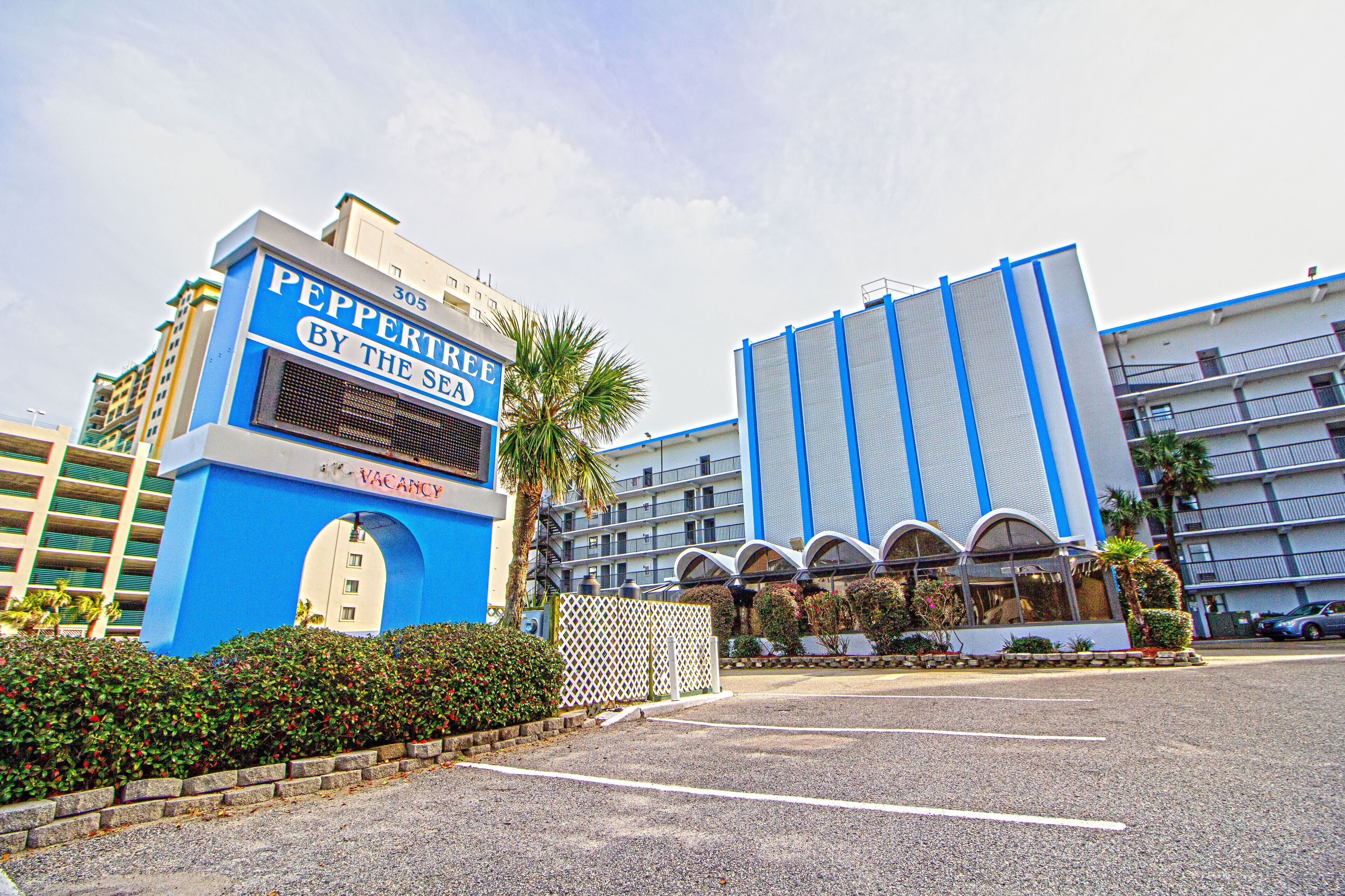 Entrada de hotel con fachada azul y blanca, un letrero que dice 'Peppertree by the Sea' y un estacionamiento al frente.