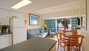 Eine kompakte Küche mit Kühlschrank, ein Essbereich mit Tisch und Stühlen, eine Couch und ein Ausblick durch eine Glastür ins Freie.