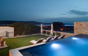 Une piscine à débordement avec des chaises longues et des parasols, un mur de pierre et une vue sur la mer et les collines.