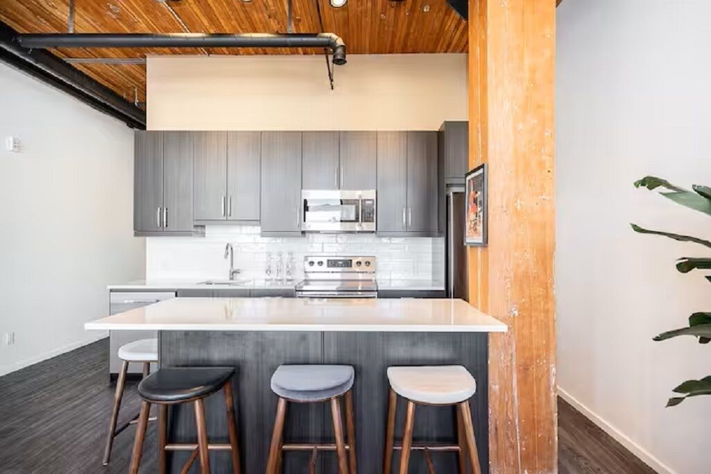 A modern kitchen with grey cabinets, stainless steel appliances, a central island with stools, and a wooden ceiling.