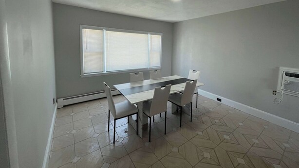 Une salle à manger avec une table rectangulaire et quatre chaises, une fenêtre avec des stores, un luminaire encastré au plafond et un climatiseur fixé au mur.