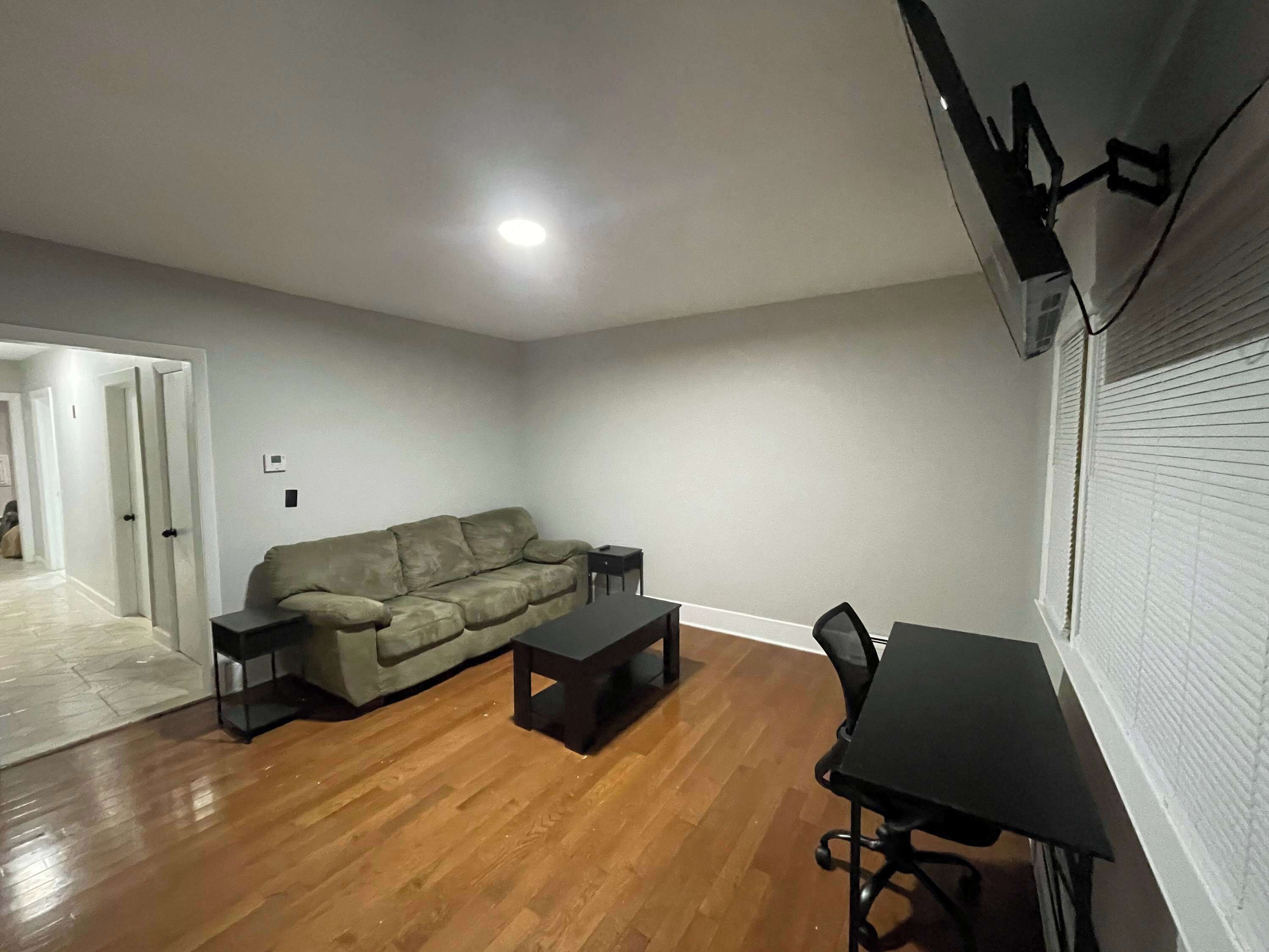 Un salon comprenant un canapé, une table basse et un bureau avec une chaise. On y trouve une porte donnant sur une autre pièce et une fenêtre avec des stores.