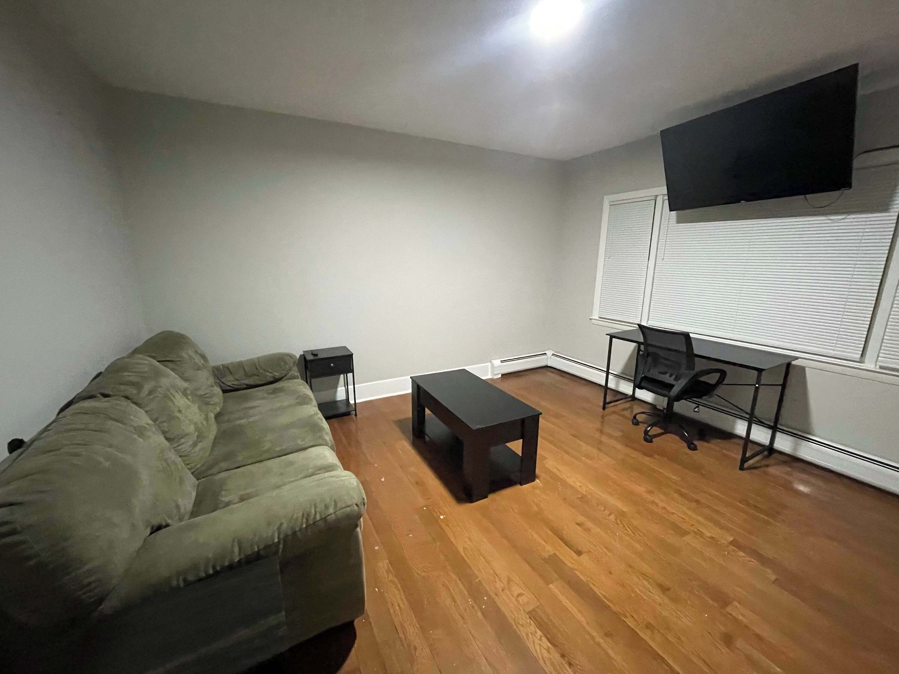 A living room with a green sofa, a small table, a TV on the wall, and a window with blinds.