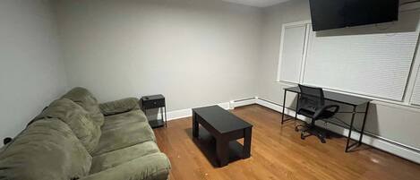 A living room with a green sofa, a small table, a TV on the wall, and a window with blinds.