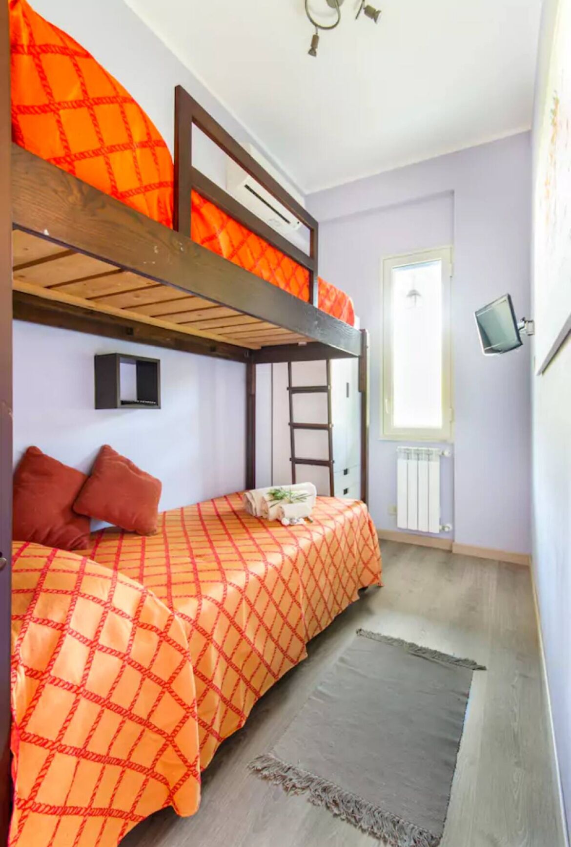 A bunk bed room with orange bedding, a small black shelf, a ladder, a television, and a door with a window.