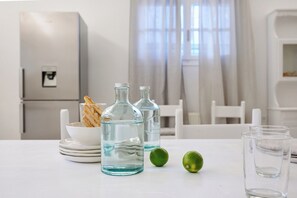 A kitchen with a refrigerator, a table with glass bottles, a bowl of breadsticks, and two limes. - Villa Afroditi by ΜΡS (Panagia)