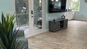 A spacious living area with a large flat-screen TV mounted on the wall, a sliding glass door leading to an outdoor balcony, and a modern kitchen with pendant lights. - Villa Nueva (Key Largo)
