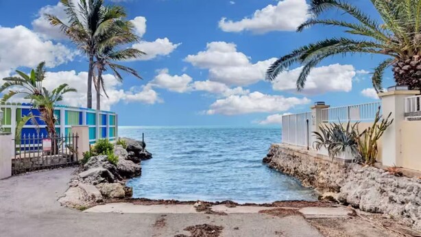 A coastal property with a paved walkway leading to a rocky shoreline, palm trees, and a clear view of the ocean. - Villa Nueva (Key Largo)